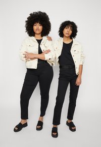 Duas pessoas vestindo jaquetas de ganga creme combinando sobre camisetas pretas, com jeans pretos e sapatos slip-on pretos, de pé lado a lado.