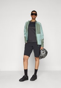 Veste zippée vert clair et gris sur un t-shirt noir, associée à un short noir et des baskets noires. Tenant un casque gris.
