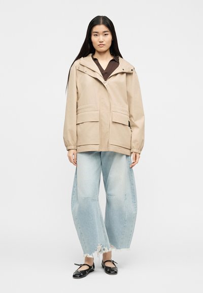 Γυναίκα που φοράει μπεζ μπουφάν με κουκούλα, ανοιχτό μπλε παντελόνι πιέτας με φθορές και μαύρες μπαλαρίνες με λουράκια στον αστράγαλο, στέκεται μπροστά σε απλό φόντο.