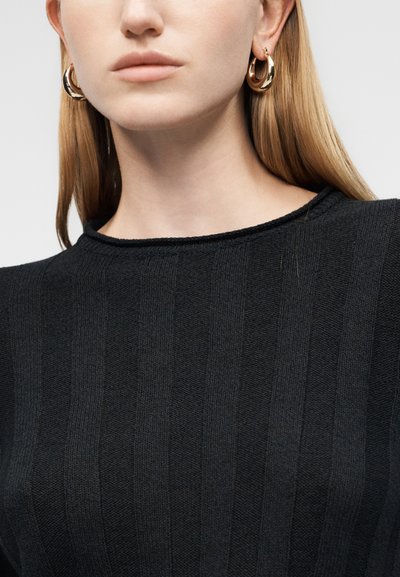 Femme aux cheveux blonds raides porte des boucles d'oreilles dorées en anneau et un pull en maille côtelée noir avec un col rond.