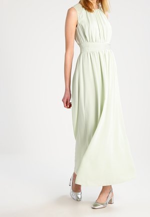 Femme portant une robe longue sans manches vert clair avec une taille cintrée et des chaussures à talons blocs argentées, se tenant devant un fond uni.