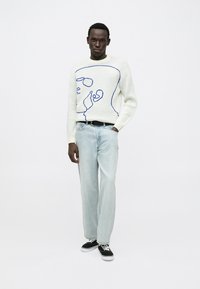 Witte gebreide trui met een blauw abstract gezicht ontwerp, gecombineerd met lichtblauwe rechte jeans en zwarte sneakers.