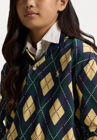 Tengeren lévő, zöld és sárga argyle mintás pólóingat fehér részletekkel. Gallért és két gombot tartalmazó gombolást kínál; anyag szerkezete szőtt.