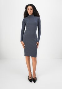 Ženska stoji v oprijetih dolgih rokavih obleki do kolen temno sivi barvi in črnih čevljih s konicami na ozadju v običajni beli barvi.