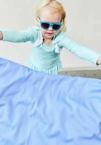 Light blue swimsuit with ruffled collar, long sleeves, and a skirted bottom. Child wearing blue sunglasses, stretching arms toward a light blue object.