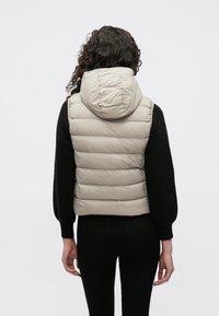 Vesta matlasată, culoare bej, cu glugă, având un design matlasat orizontal; combinată cu un top negru cu mâneci lungi; stil minimalist.