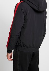 Svart huva jacka med röda sidostripe, elastisk midja och mudd. Slät textur med en lätt design, med ett sportigt utseende.