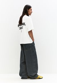 Hvid oversized t-shirt med sort tekst på ryggen, parret med løstsiddende mørke denim jeans og sko med gule akcenter. Glat stof tekstur.