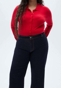 Femme portant un cardigan rouge ajusté boutonné et un jean bleu foncé taille haute, debout avec les mains derrière le dos.