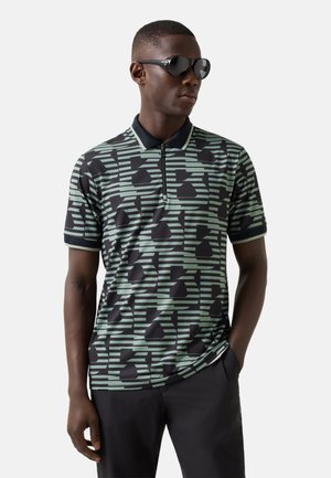 Man met een zwarte zonnebril en een zwart-groen polo met geometrisch patroon en korte mouwen, met een zwarte broek, staand met een hand in zijn zak.