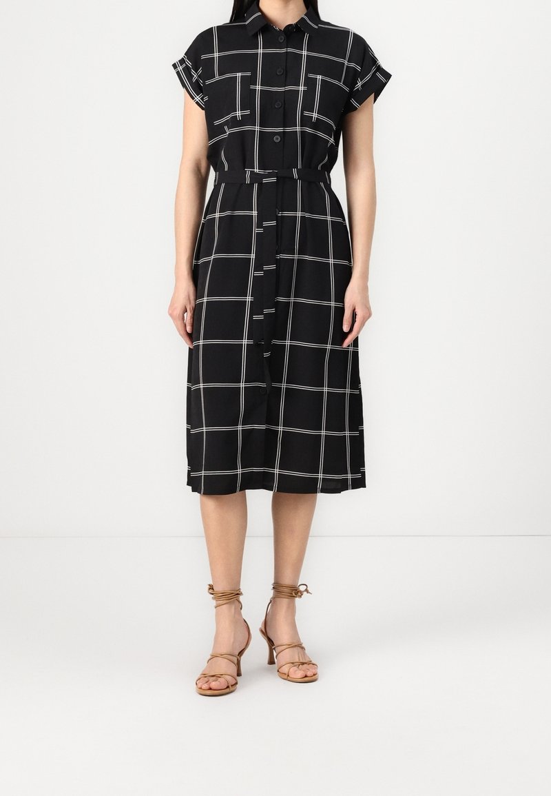 Femme portant une robe chemise midi noire avec un motif à carreaux blancs, boutonnée à l'avant, manches courtes, ceinture et talons ouverts beiges.