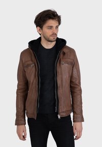 Giacca di pelle marrone con finitura testurizzata, chiusura con zip frontale, due tasche sul petto e cappuccio foderato in felpa nera. Il modello indossa un top nero e jeans.