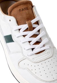Baskets en cuir blanc avec une languette en suède marron, présentant des accents verts, des lacets blancs, des détails perforés sur le bout, et une semelle en caoutchouc.