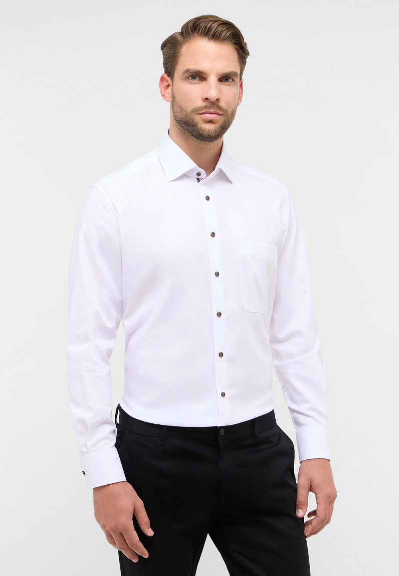 Camicia bianca a maniche lunghe con colletto button-down, bottoni marroni e taschino sul petto. Il tessuto appare liscio con un design aderente.