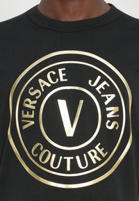 Tricou negru cu un logo circular mare de culoare aurie, având textul "Versace Jeans Couture" și un "V" central. Material moale, mâneci scurte.