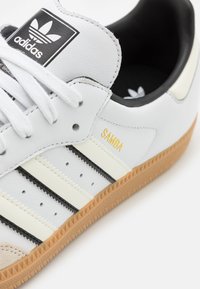 Vita lädersneaker med svarta accenter, tre svarta ränder och en gummisula. Har "Samba" i guld på sidan. Texturerad tånät.