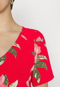 Vestido floral vermelho com decote em V, mangas curtas e flores vibrantes em rosa e verde num tecido texturizado.