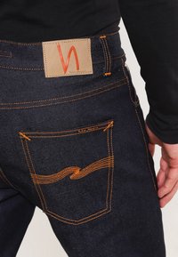 Mörka denimjeans med orange sömmar, en läderetikett med ett stiliserat 'W' och en böjd fickdesign.