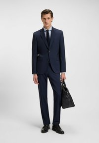 Marineblauw pak met een aansluitende blazer en rechte broek, gecombineerd met een lichtblauw shirt en een donkere stropdas. Houdt een zwart gestructureerde tas vast.