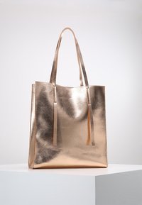 Metaliczna torba tote w kolorze różowego złota, gładka faktura, dwie długie rączki, otwarty górny z pasującymi frędzlami, prostokątny kształt, minimalistyczny design.
