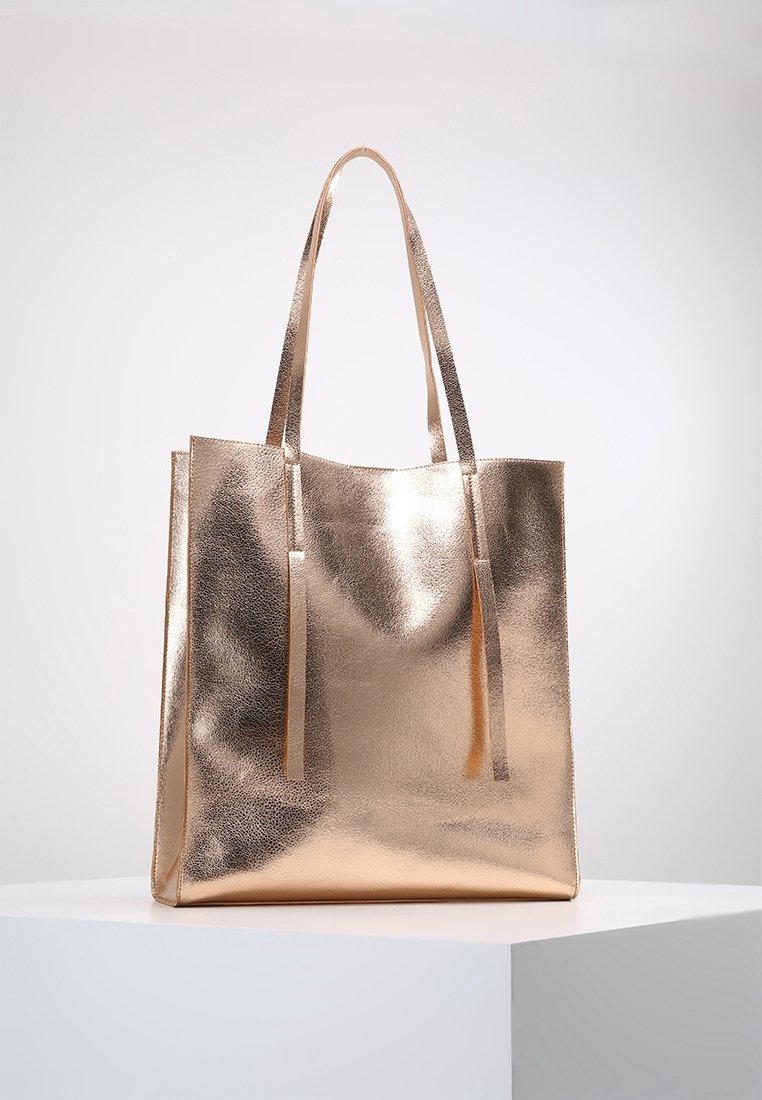 Metaliczna torba tote w kolorze różowego złota, gładka faktura, dwie długie rączki, otwarty górny z pasującymi frędzlami, prostokątny kształt, minimalistyczny design.