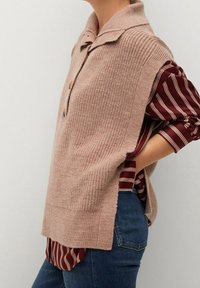 Stickad beige tröja med krage, som visar upp ett strukturerat mönster och två knappar. Kombinerad med en randig burgunderfärgad skjorta under.