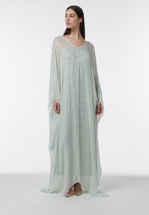 Vrouw draagt een vloeiende, halfdoorzichtige lichtgroene jurk met lange mouwen, staat en kijkt naar haar linkerzijde tegen een witte achtergrond.