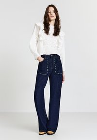 Blusa bianca con rouches e dettagli in pizzo, abbinata a jeans a gamba larga blu scuro con borchie decorative lungo le tasche.