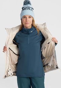 Pull bleu à fermeture éclair, porté sous une veste crème au intérieur lisse. Le mannequin porte un bonnet bleu clair avec un pompon.