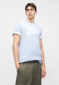 Νεαρός άνδρας που φοράει ανοιχτό μπλε μπλουζάκι Nike και ελιάς πράσινο παντελόνι, στέκεται με τα χέρια στις τσέπες, κοιτάζοντας προς τα πλάγια σε απλό λευκό φόντο.