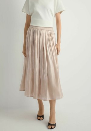 Kobieta ubrana w jasnobeżową plisowaną spódnicę midi, biały top z krótkim rękawem i czarne buty na obcasie z odkrytymi palcami, stojąca na jednolitym tle.