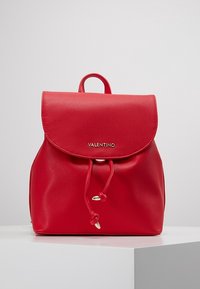 Mochila de cuero sintético rojo con cierre de solapa, detalle de cordón y herrajes dorados. Superficie texturizada y logo en la parte frontal.