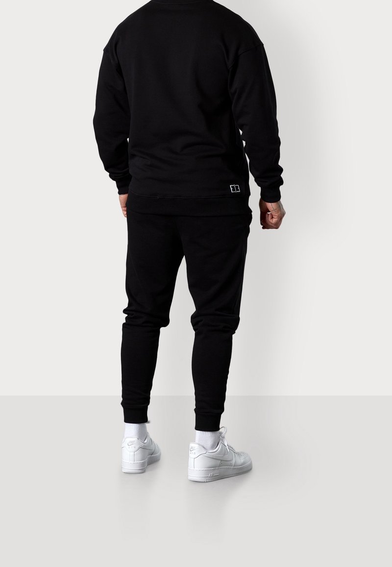 Personne portant un pantalon de survêtement noir et un sweat-shirt associés à des baskets blanches Nike Air, debout face à un mur blanc uni.