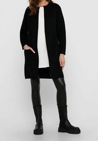 Černý dlouhý kabát z měkké tkaniny, s kulatým výstřihem, otevřeným předním dílem a dvěma bočními kapsami. Nositelný přes bílou košili a černé kalhoty.