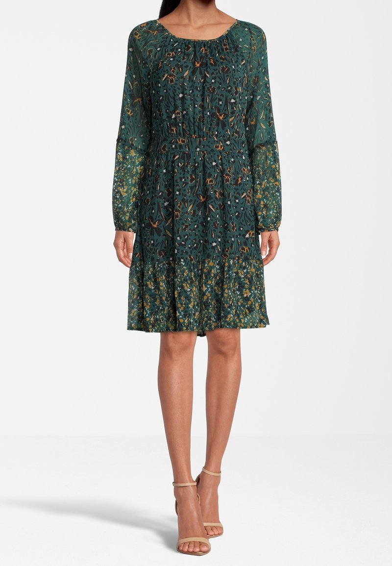 Femme portant une robe longue à fleurs verte avec une taille cintrée et des talons beiges à bride à la cheville, debout devant un fond uni.