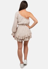 Hellbeige Ein-Schulter-Kleid mit Rüschensaum, gerafftem Taillenbereich und strukturiertem Stoff. Kombiniert mit weißen Sneakers.