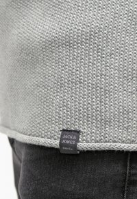 Světle šedý pletený svetr s lemem a černou značkovou etiketou "JACK & JONES ONM Co." na černých džínách.