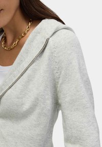 Hellgraue Zip-Hoodie mit weicher Textur, figurbetontem Design und gerippten Akzenten. Verfügt über einen Frontreißverschluss und eine Kapuze. Sichtbare goldene Kettenhalskette.