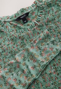 Green floral blouse featuring smocked texture, gathered neckline, and short sleeves with a floral pattern. Fabric is light and textured.