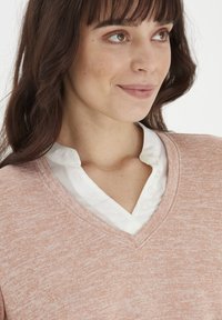 Rosa sweater med V-hals over en hvid skjorte med krave, der har en blød tekstur og et subtilt meleret mønster.