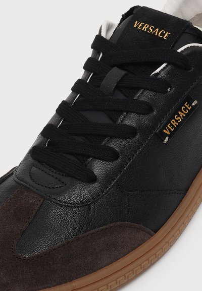 Baskets noires Versace avec lacets noirs, logo Versace doré, bout en daim marron et semelle en caoutchouc marron à motif clé grec.