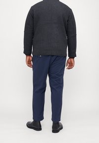 Grau-wollene Jacke mit hohem Kragen, kombiniert mit navyblauen Hosen und schwarzen Stiefeln. Die Jacke hat eine taillierte Passform und strukturierte Schultern.