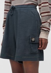Shorts texturés bleus avec poche cargo arborant un patch logo "Santa Cruz", portés avec un haut beige à manches longues avec des rayures bordeaux.