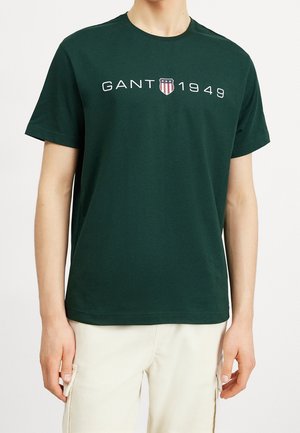 Person iført mørkegrøn kortærmet T-shirt med "GANT 1949" og skjoldlogo på brystet, kombineret med off-white bukser.