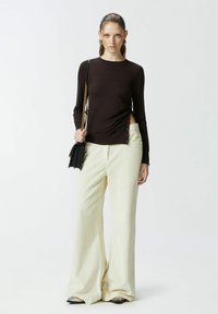 Top a maniche lunghe marrone scuro con dettagli a bottoni laterali, abbinato a pantaloni larghi giallo chiaro e una borsa a spalla nera con tracolla a catena.