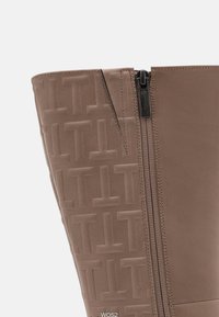 Bota de rodilla color topo con patrón en relieve de "T" en el lado izquierdo, cuero liso en el derecho y una cremallera con tira negra en el centro.