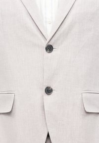 Ανοιχτό μπεζ σακάκι με σχέδιο ψαροκόκκαλο, με δύο σκούρα κουμπιά κλεισμένα, λευκό πουκάμισο με διακριτικές κάθετες ρίγες από κάτω.