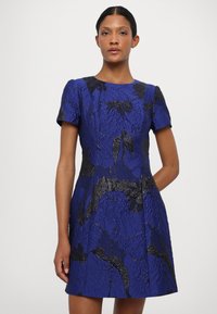 Īss zils kleita ar teksturētu audumu, kas rotāts ar melnu ziedu rakstu, īsām piedurknēm, apaļu dekoltēju un pieguļošu siluetu.