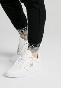 Vita tennis med läder och guldaccent, med en texturerad sula och logodetalj. Bärs med svarta avsmalnande byxor med märkta manschetter.