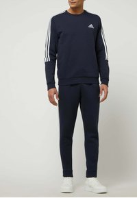 Felpa navy e pantaloni jogger abbinati, entrambi in morbido tessuto, presentano accenti a tre strisce bianche sulle maniche e un logo sul petto. Sneakers bianche completano il look.